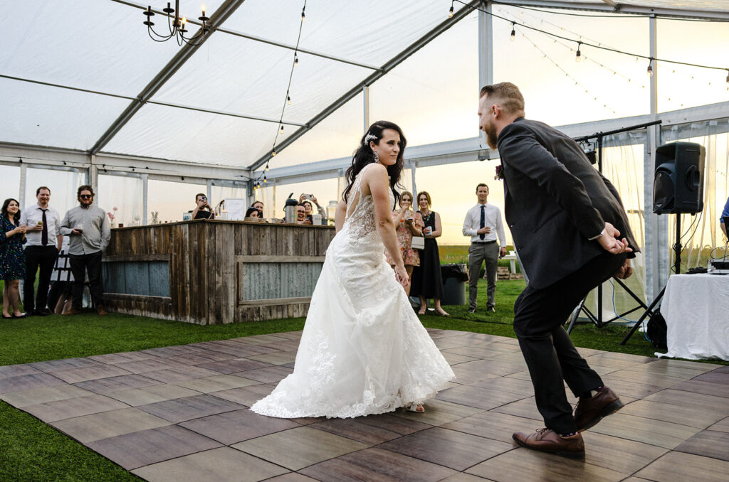 Bride and groom share a dance on a wooden floor at an outdoor wedding reception, with guests looking on.