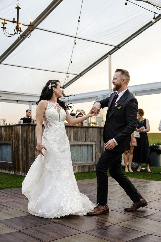 A bride and groom share a joyful dance outdoors under a tent with guests looking on.