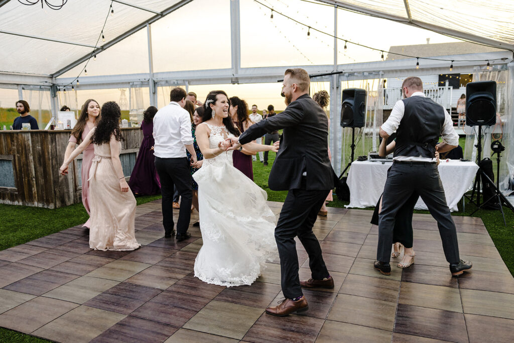 Couple dancing on a wooden floor at an outdoor wedding reception with guests watching and a dj in the background.