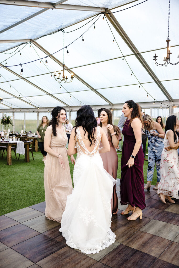 A bride in a lace wedding dress converses with guests at an outdoor reception under a tent adorned with string lights.
