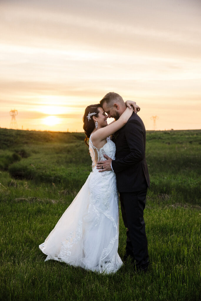 A couple in wedding attire embraces in a field at sunset.
