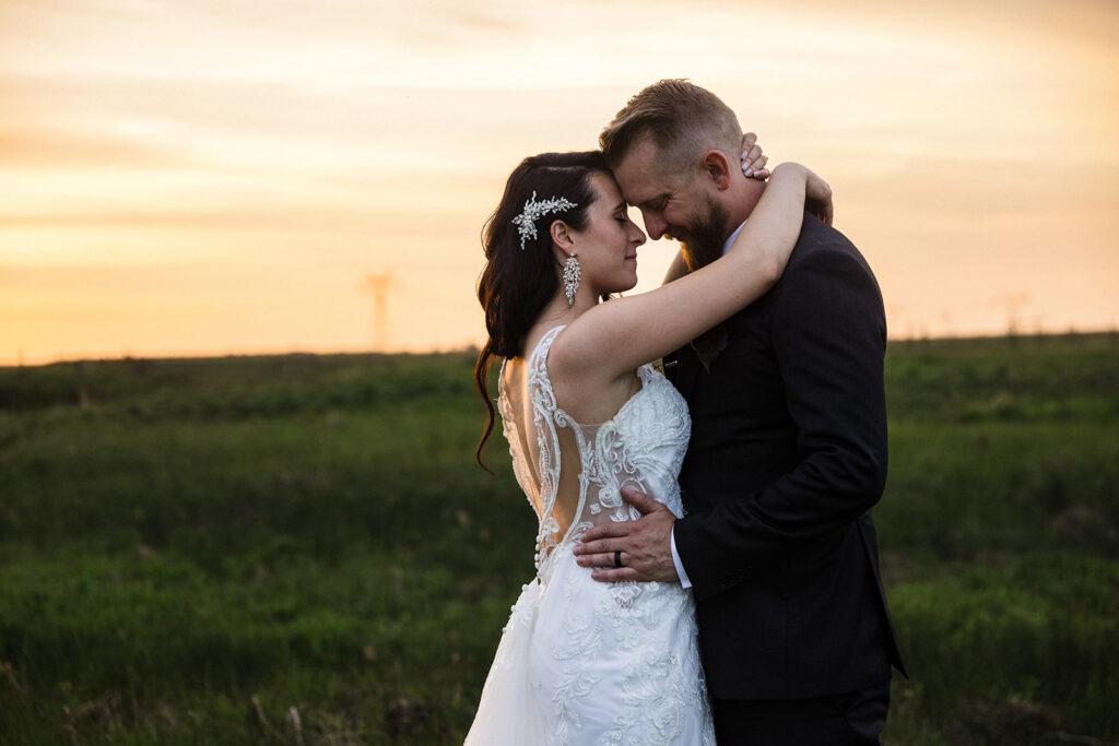 A couple in wedding attire embracing at sunset in a field.