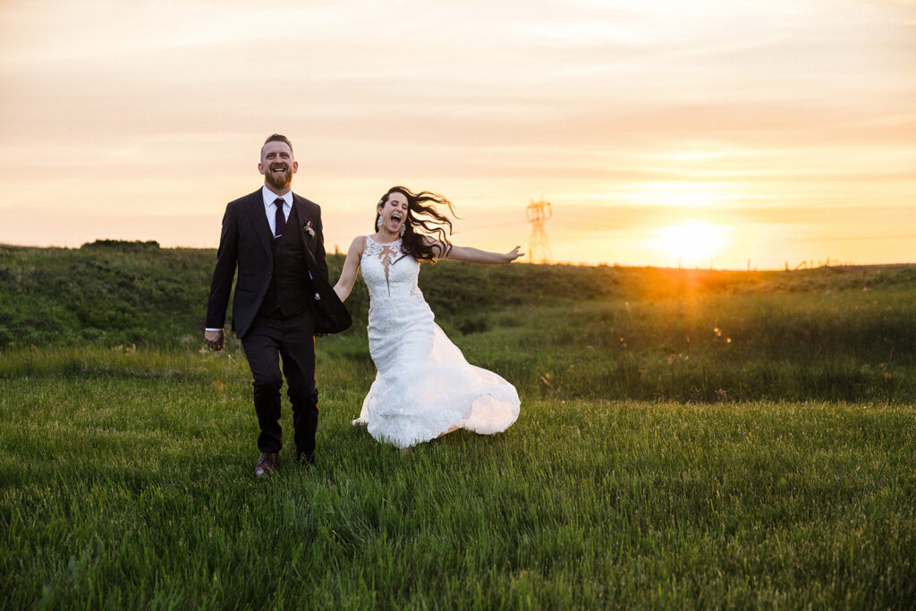 A joyful bride and groom running through a field at sunset.