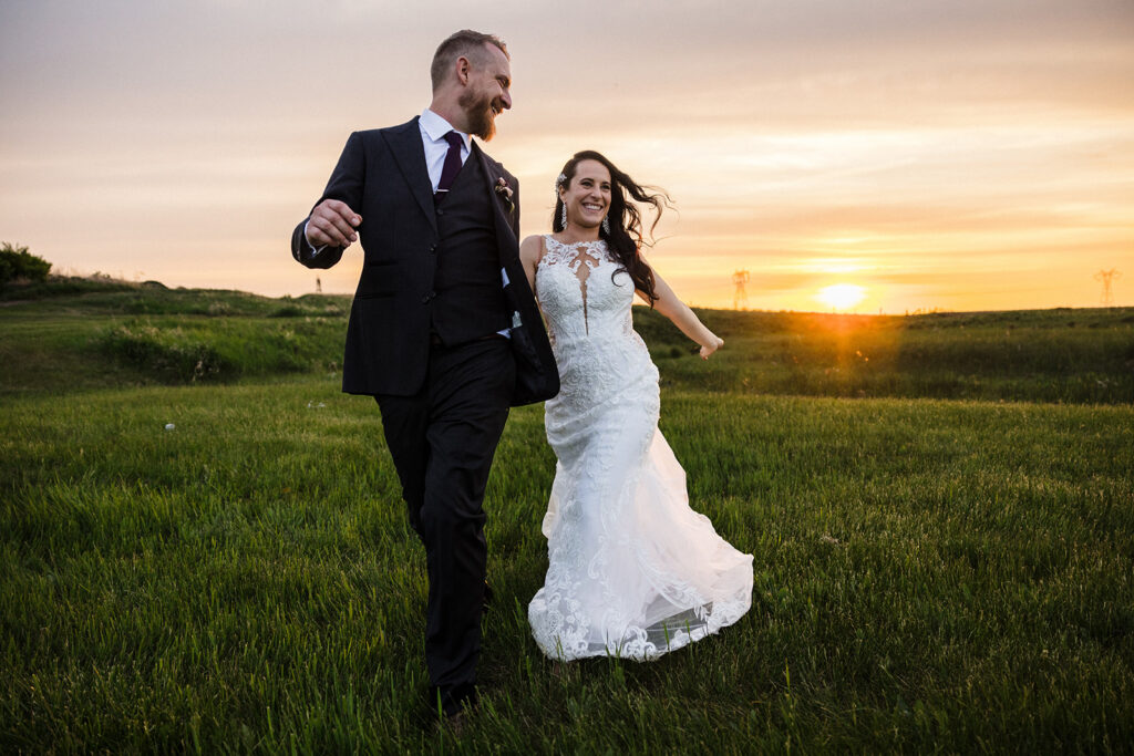 Bride and groom walking hand in hand through a field at sunset.