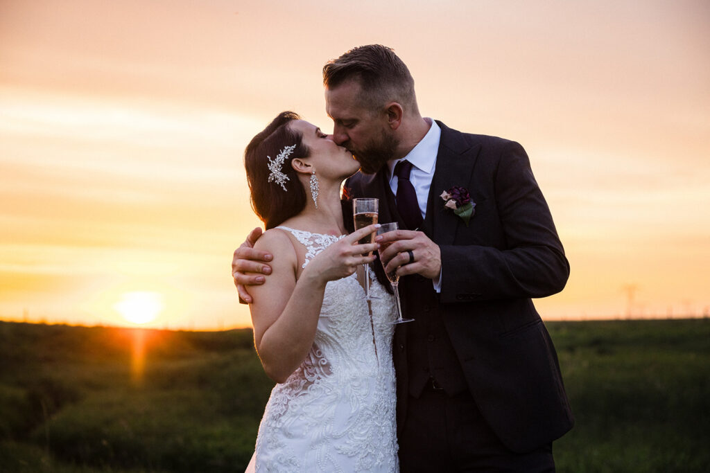 Bride and groom sharing a kiss while holding champagne glasses at sunset.