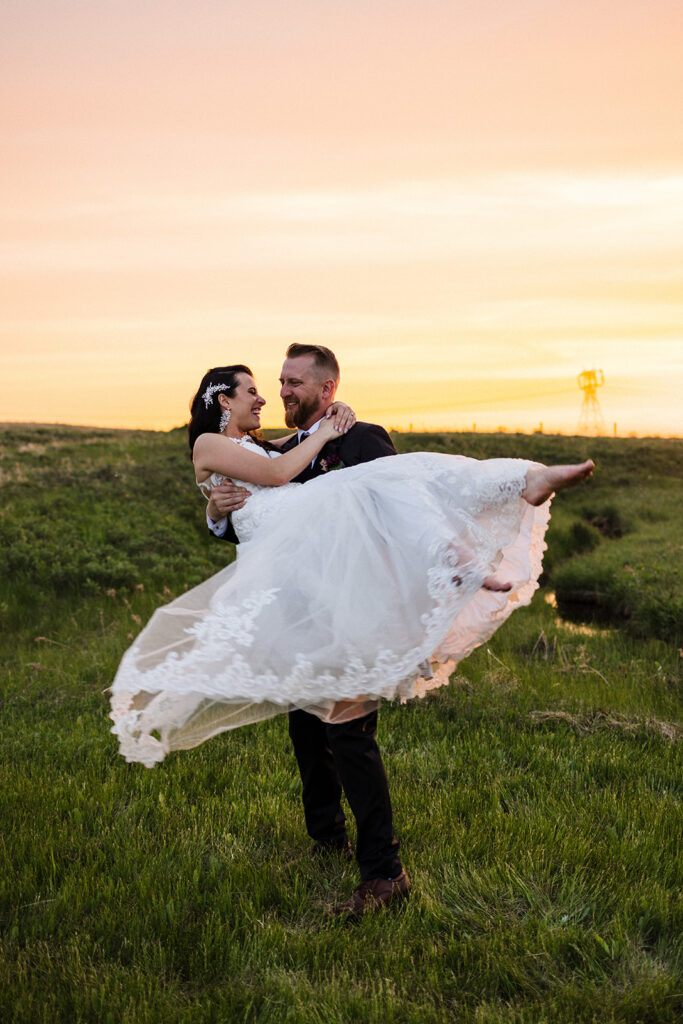 Groom lifting bride in a joyful pose against a sunset backdrop in a grassy field.