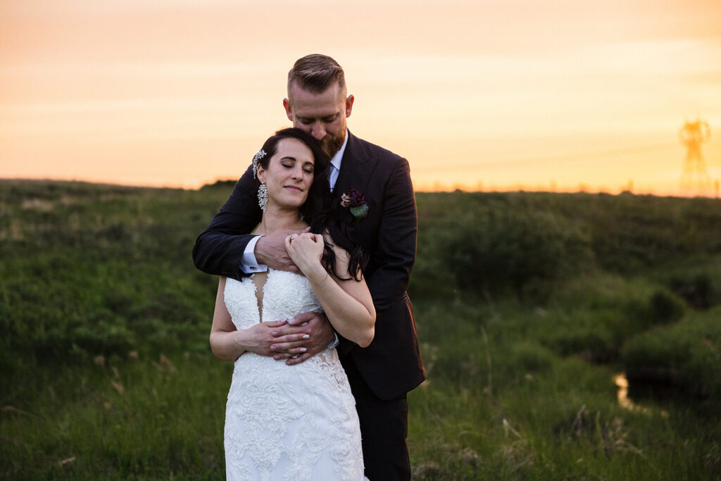 A couple in wedding attire embracing in a field at sunset.