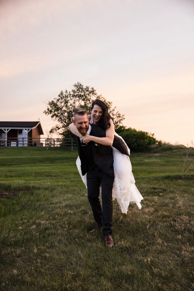 Groom carrying bride on his back in a grassy field at dusk.