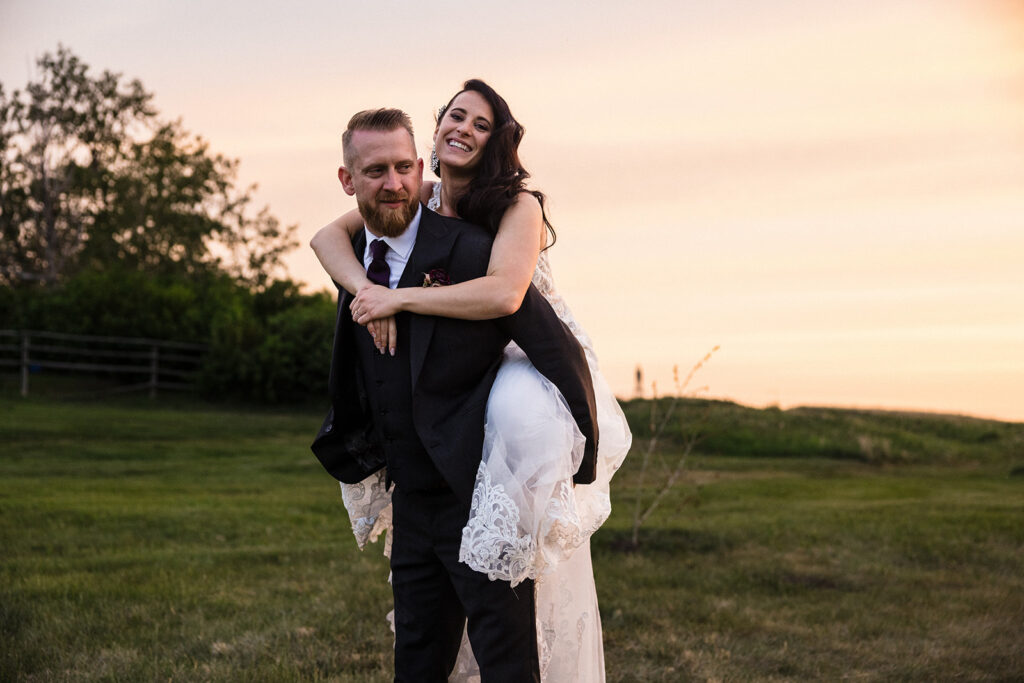 A bride with a piggyback ride on the groom, both smiling in a field at sunset.