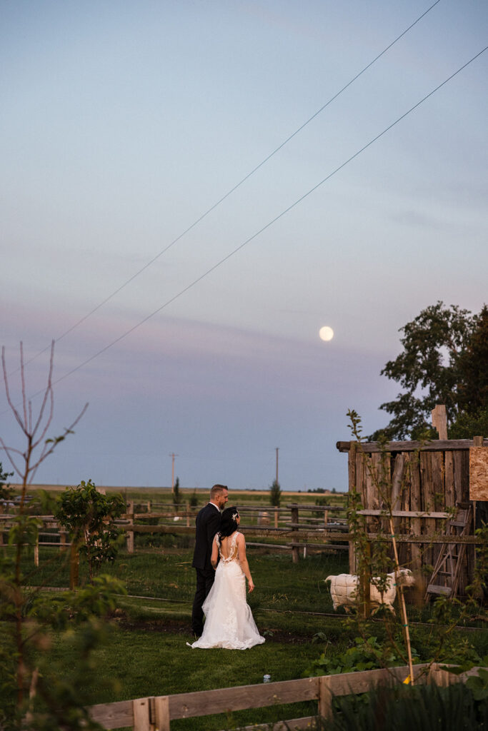 A couple in wedding attire sharing a moment in a rustic farm setting under a moonlit sky.